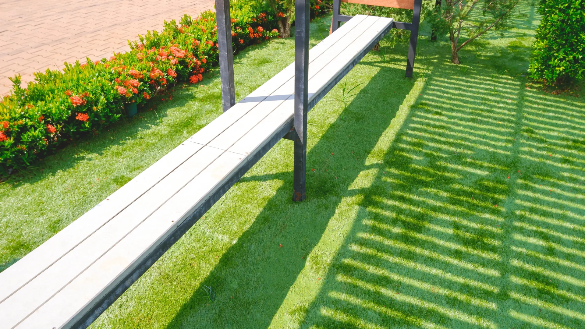 Artificial turf lawn with a long slatted bench, a flowering hedge, and striped shadows from a pergola