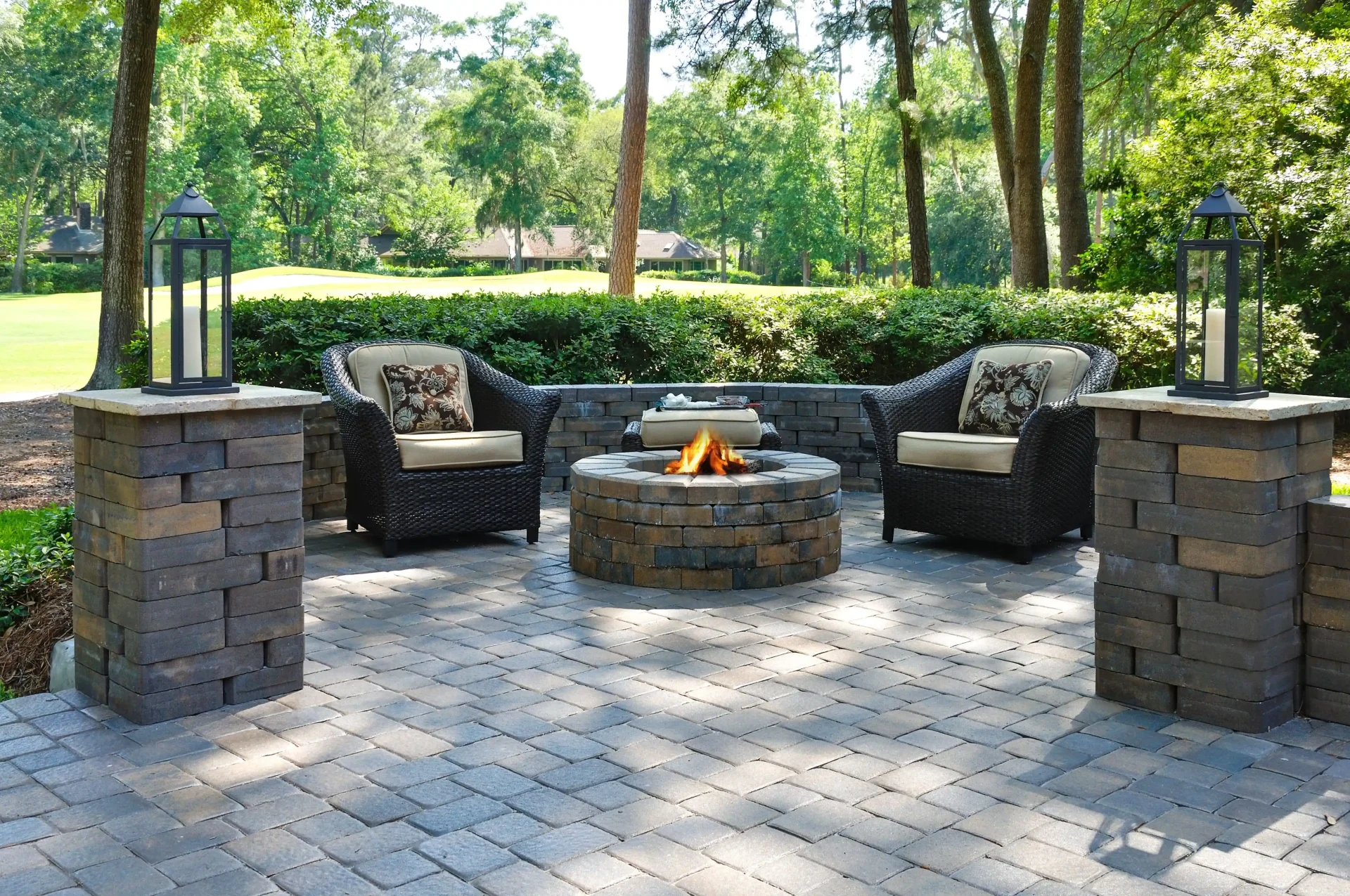 Paver patio with a round stone fire pit, curved seat wall, wicker armchairs, and lantern-topped pillars